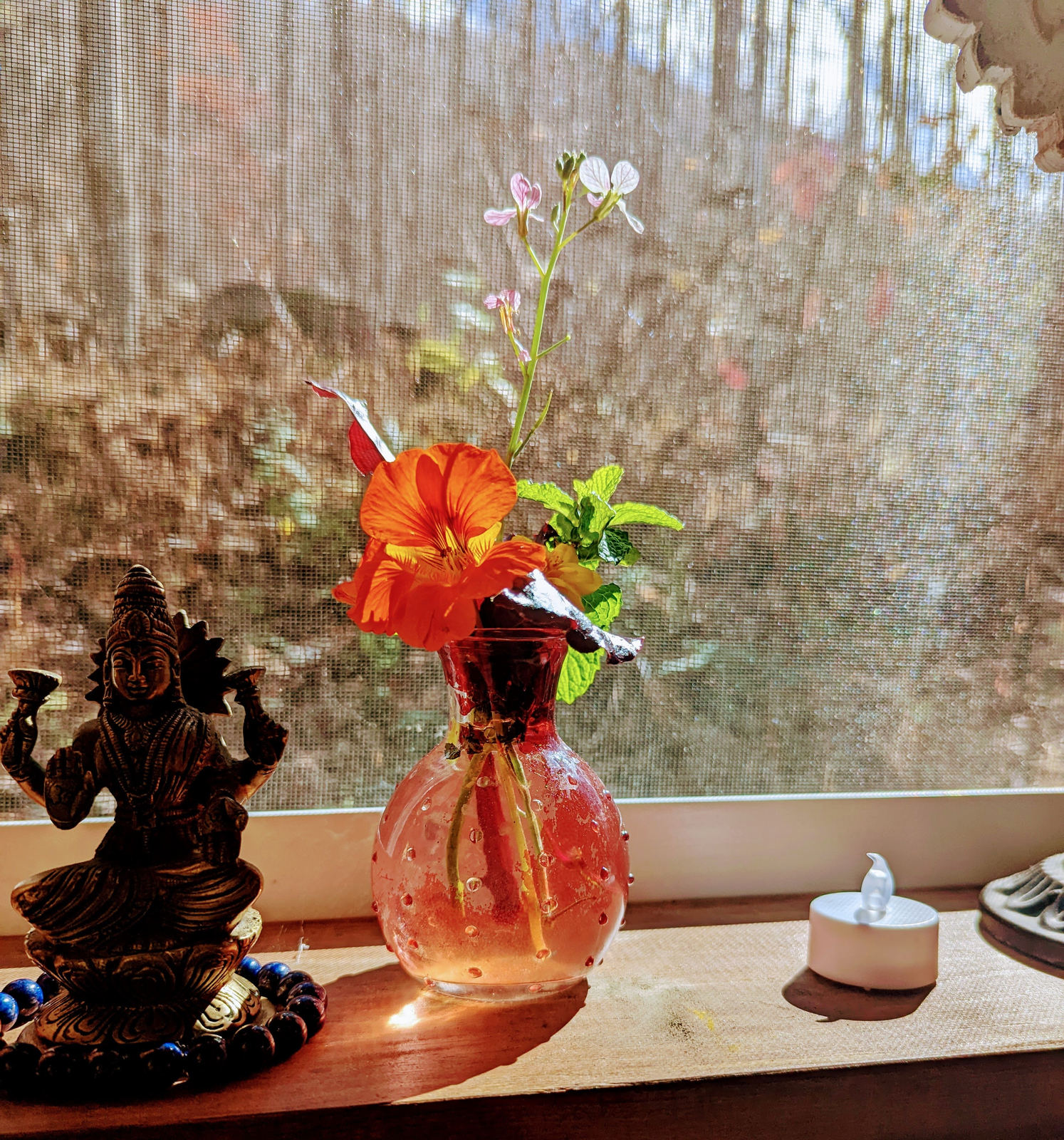A small altar by a window with a bronze figure, orange lilies in a glass vase, and a candle